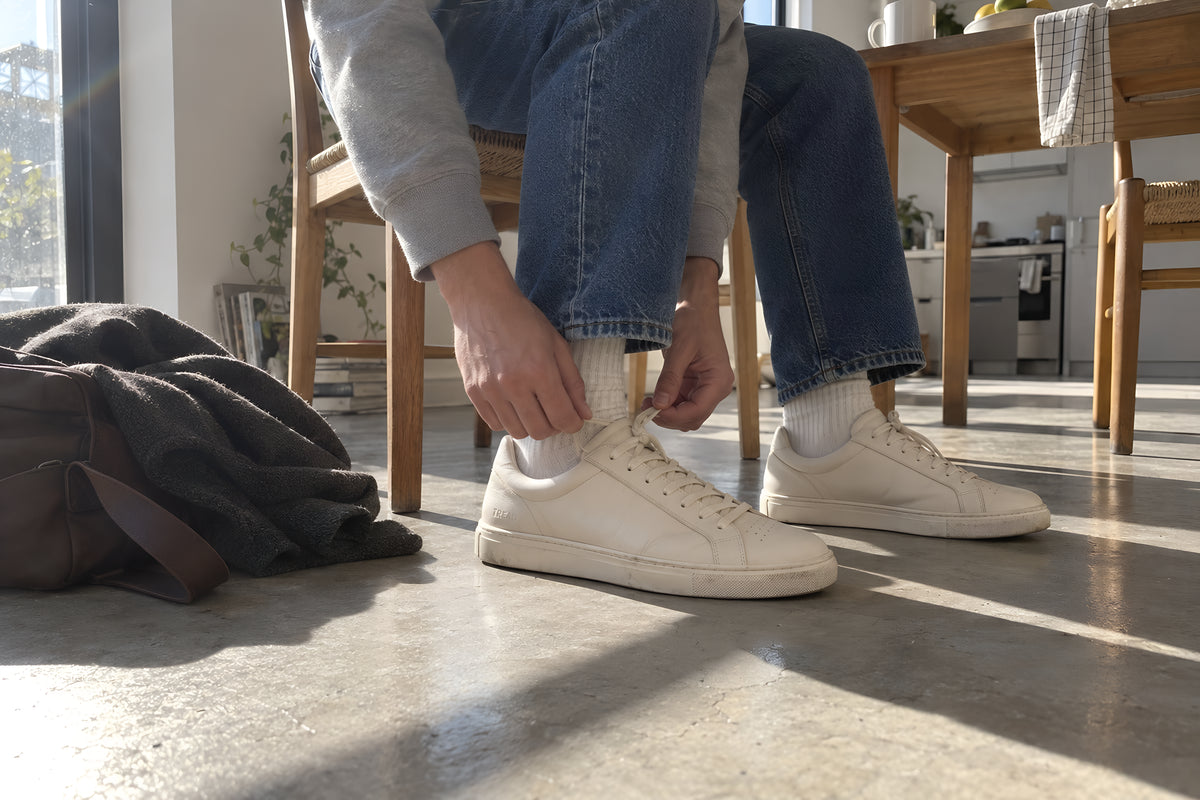 Someone perched on the edge of a kitchen chair pulling the laces tight on a bone TREAD Court sneaker, only the legs and hands in frame, a bag and a coat dumped on the floor beside them. Anatomy matters here: one left foot and one right foot, correctly mirrored, the laced shoe on the near foot and the other flat on the floor. Daytime, hard sun, no candles lit.