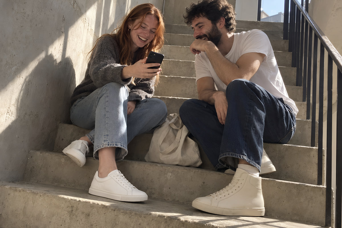 A woman with red hair in white TREAD Court sneakers and a man with a dark beard in bone TREAD Court High sitting together on a concrete stairwell, both laughing at something on her phone which she is holding out for him to see, a bag between them. Warm and easy, not an argument. Daytime, hard sun down the stairwell, no candles lit.