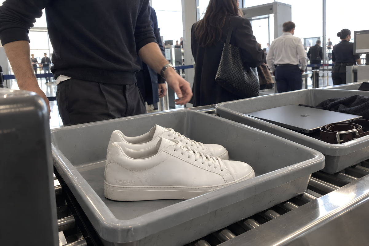 A pair of white TREAD Court sneakers in a gray security tray on the belt at an airport, a laptop and a belt in the next tray along. Strip light, no candles lit.