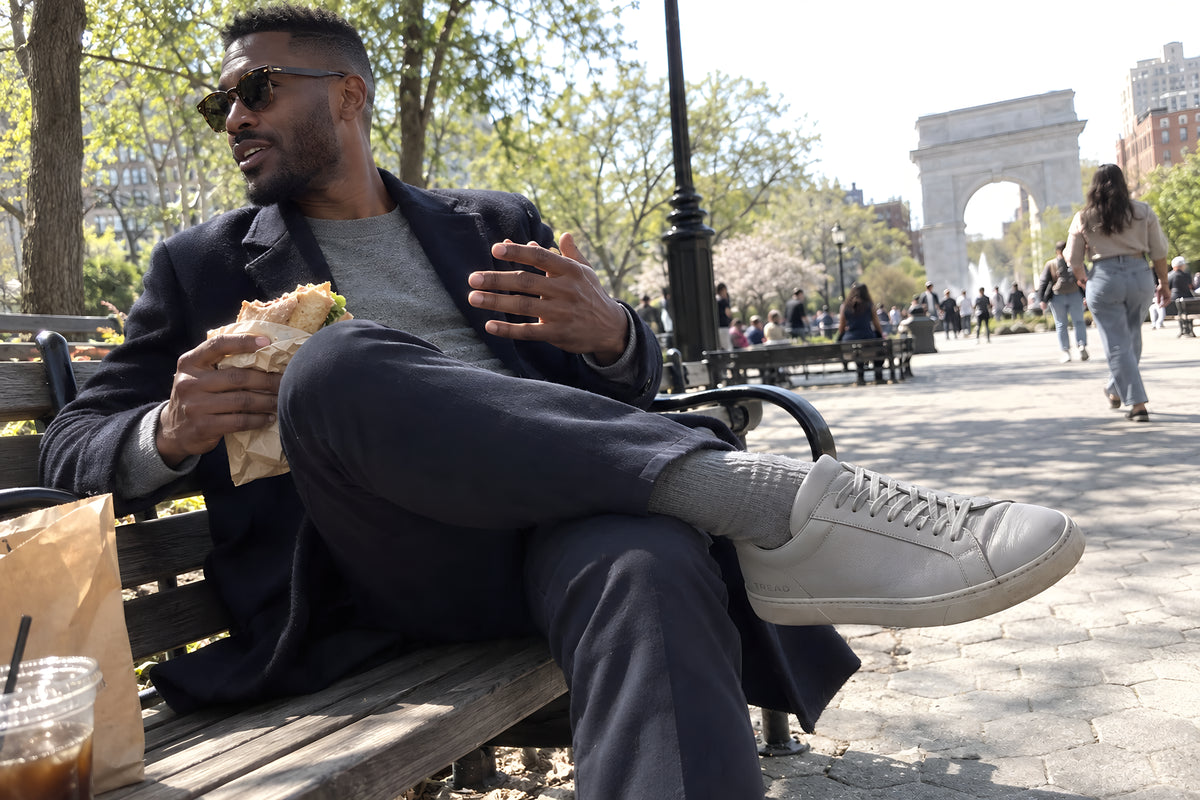 A good-looking man in his thirties sitting back on a park bench with an ankle across his knee, gray TREAD Court sneaker up, sandwich in his other hand. Daytime, hard sun, no candles lit.