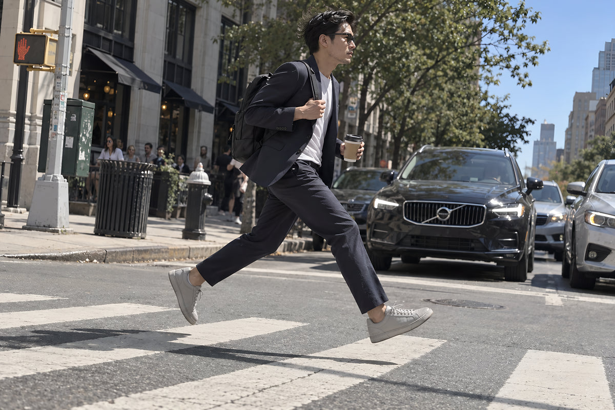 A man striding across a zebra crossing in gray TREAD Court sneakers, caught with both feet off the ground, traffic waiting. Daytime, hard sun, no candles lit.