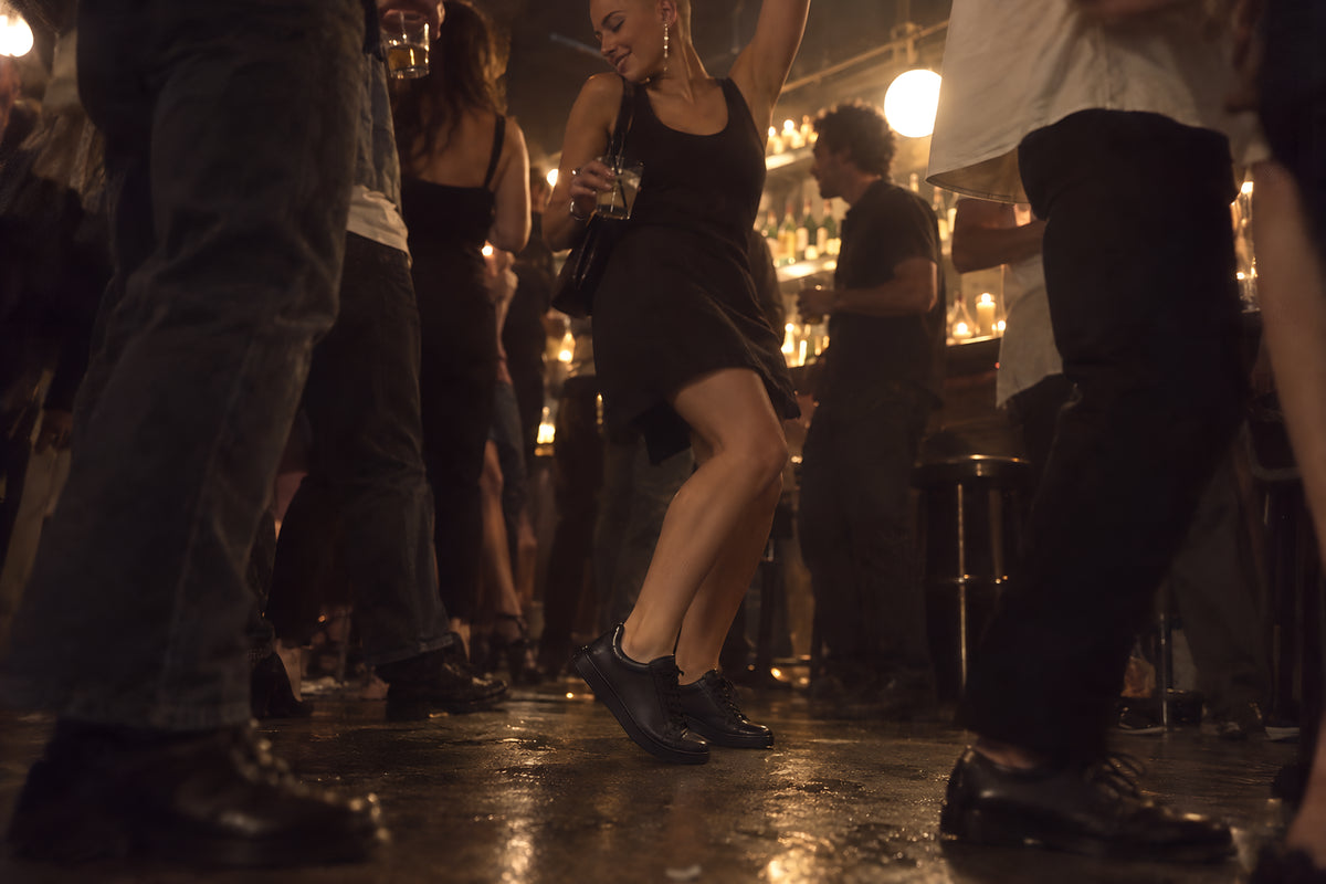 Feet and shins on a busy bar floor late at night, a woman in black TREAD Court sneakers up on the balls of her feet dancing, other legs crowded round.