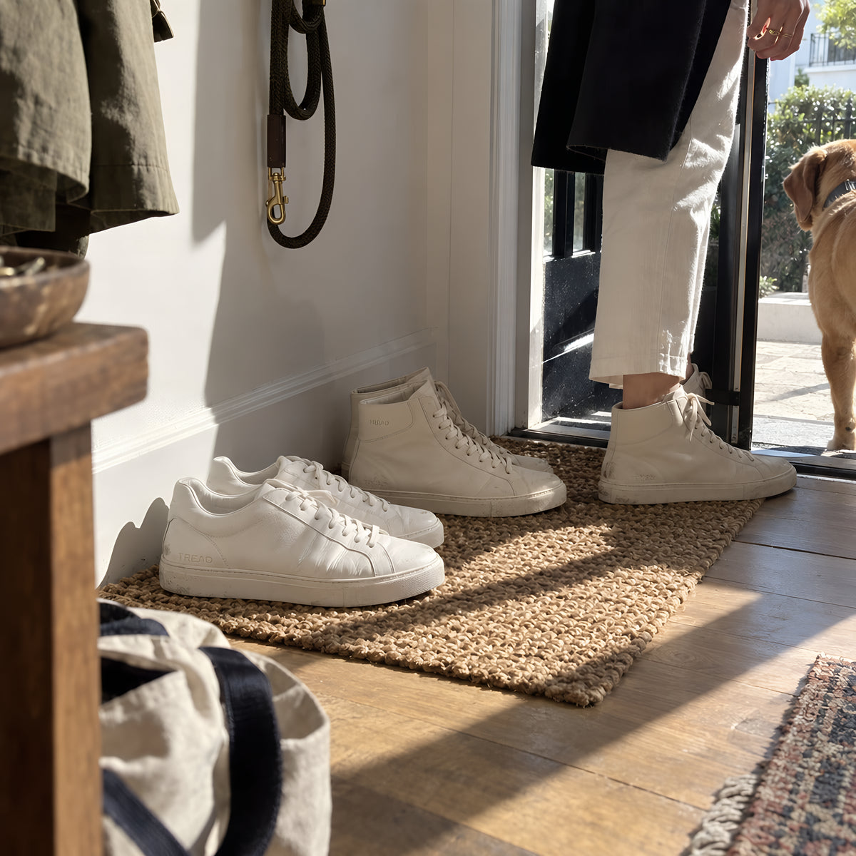 Two pairs of TREAD Courts lined up inside a front door on a mat, one white and one bone high top, a dog lead hanging above them. Daytime, hard sun, no candles lit.