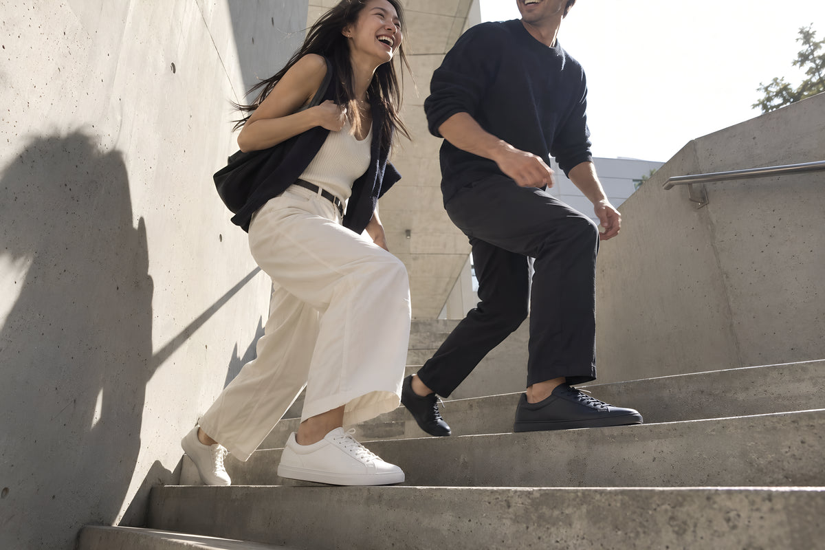 A couple in their late twenties taking the steps of a concrete stairwell two at a time, one in white TREAD Court and one in navy, both laughing at something ahead of them. Daytime, hard sun, no candles lit.