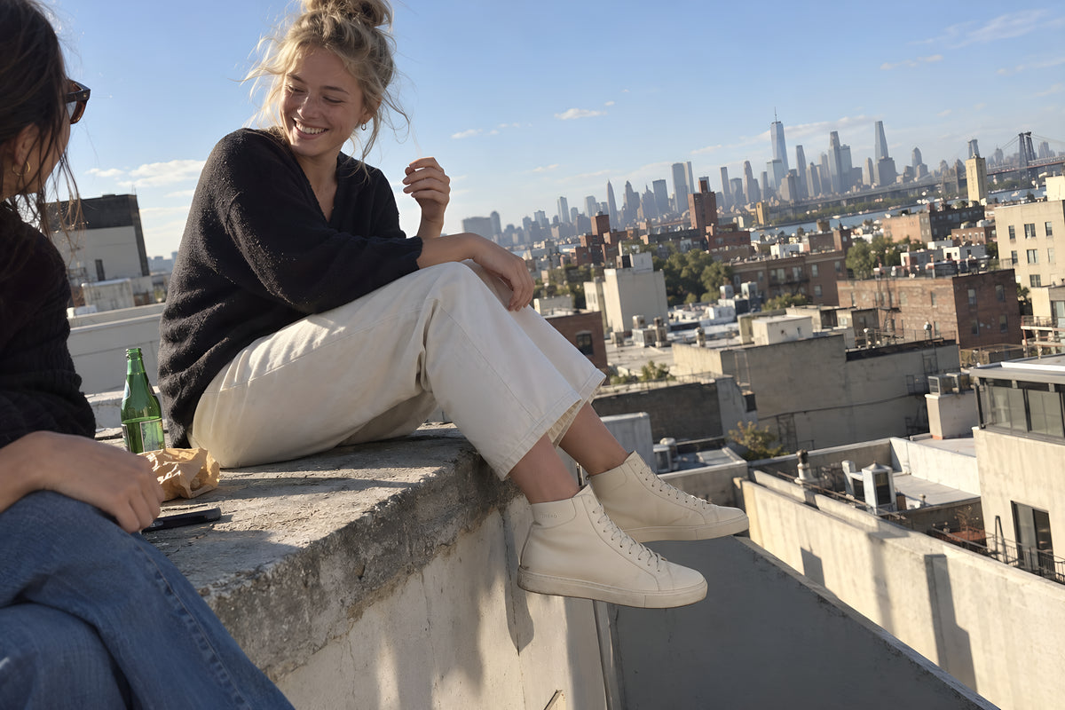 Two friends sitting on the edge of a roof terrace with their legs hanging, one in bone TREAD Court High, city stretched out beyond. Daytime, low hard sun, no candles lit.