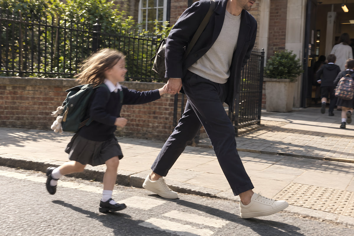 A man walking fast on a school run in bone TREAD Court sneakers with a small child half running to keep up holding his hand, book bag swinging. Daytime, hard sun, no candles lit.