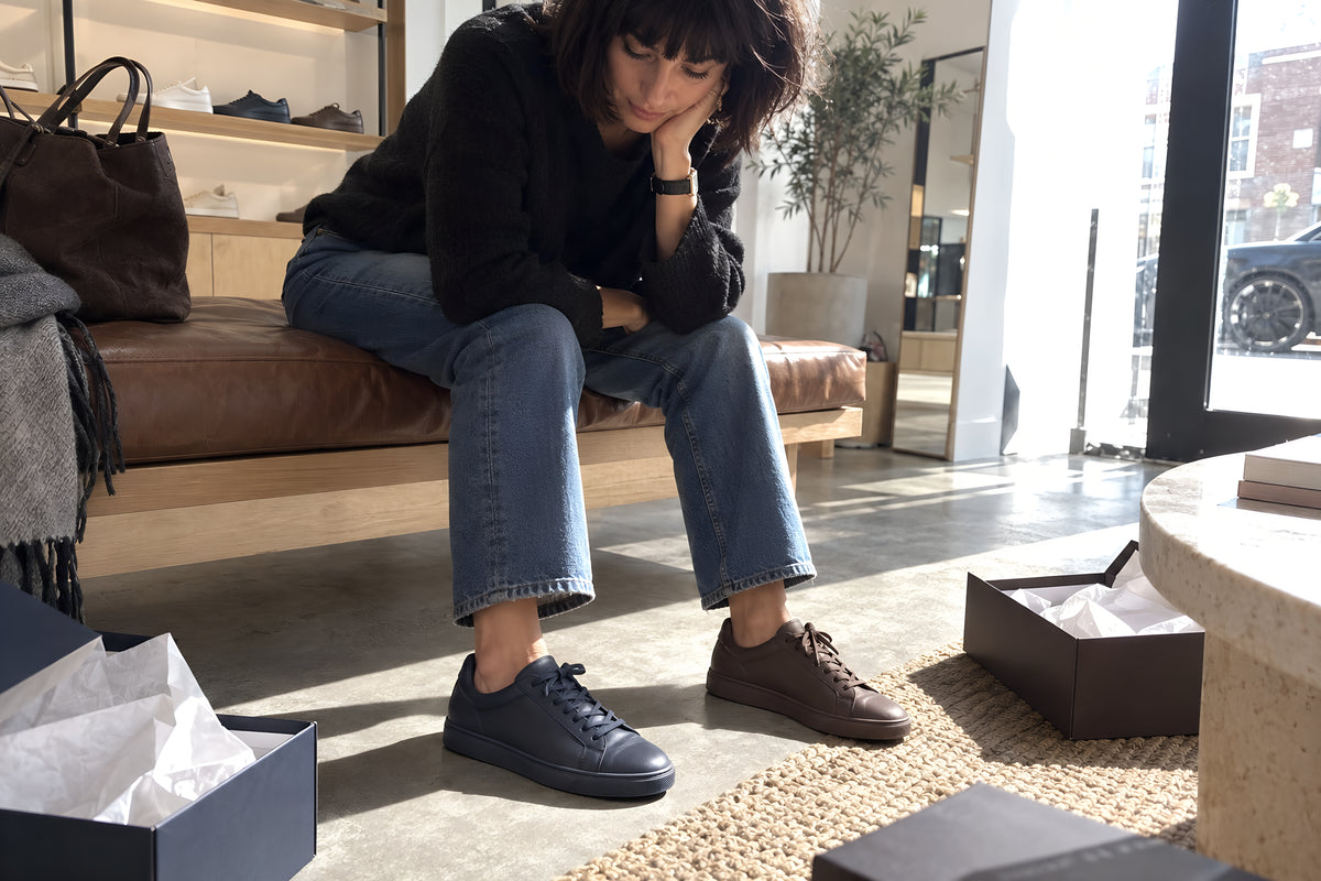 Someone sitting on a shop bench with one navy TREAD Court on and one chocolate on, looking down at both feet deciding, boxes open around them. Daytime, hard sun, no candles lit.