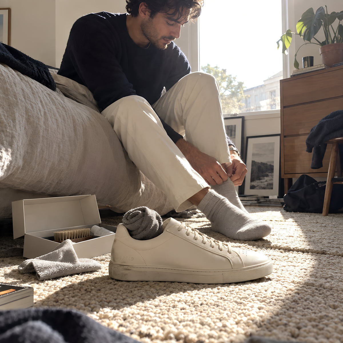 A bone TREAD Court on a bedroom floor beside an open TREAD Care Kit, brush and cloth out, a rolled pair of socks tucked in the shoe. Daytime, hard sun, no candles lit.