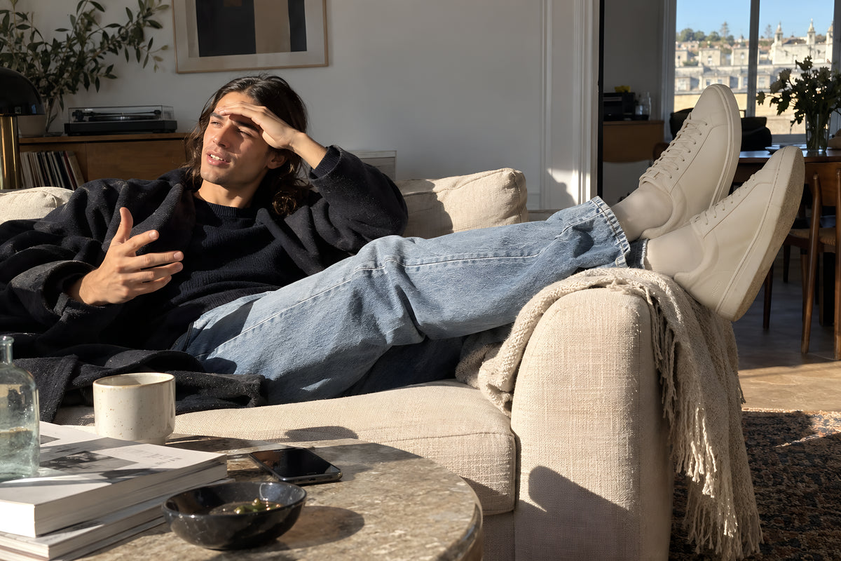 Someone come in and flopped onto a sofa with bone TREAD Court sneakers still on and feet up over the arm, coat still half on. Daytime, hard sun, no candles lit.