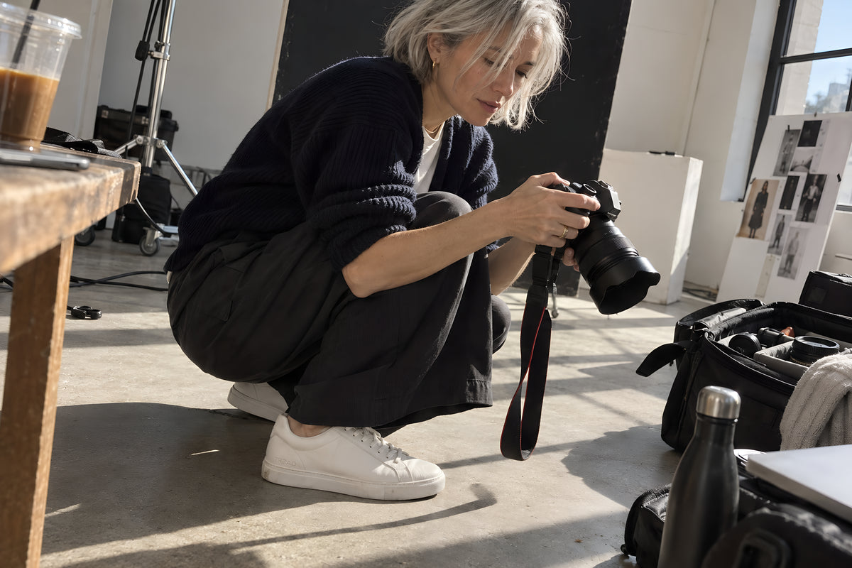 A photographer crouched on a studio floor in white TREAD Court sneakers checking the back of a camera, gear bags open around her. Daytime, hard window light, no candles lit.