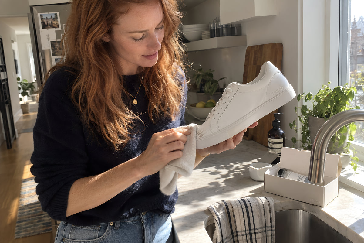 A woman at a kitchen sink wiping a scuff off the toe of a white TREAD Court with a cloth from the TREAD Care Kit, the shoe held up to the light to check. Daytime, hard sun, no candles lit.